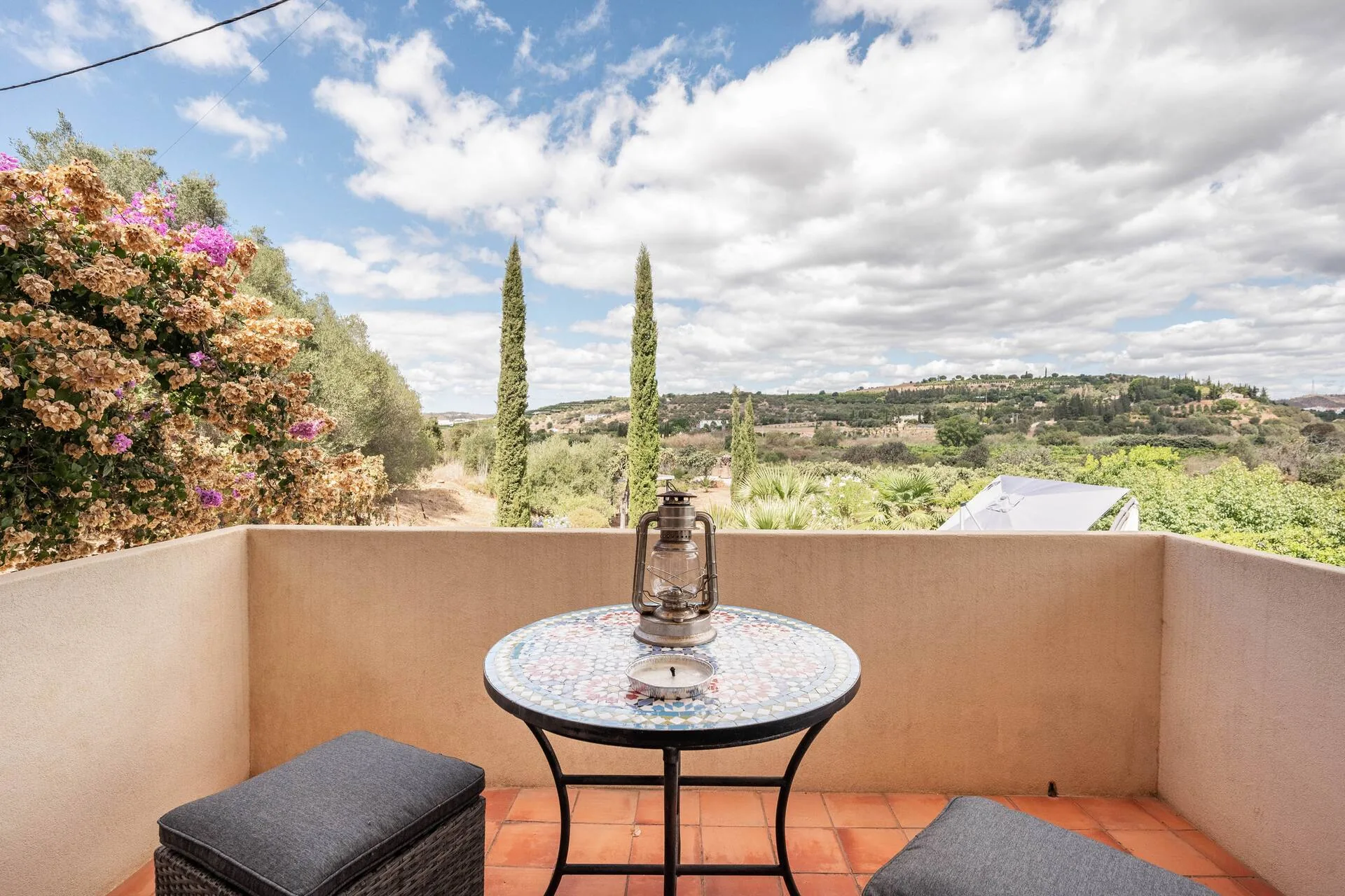 A small private balcony with a mosaic bistro table and lantern, framed by bougainvillea, looking out over the cypress trees and hills.