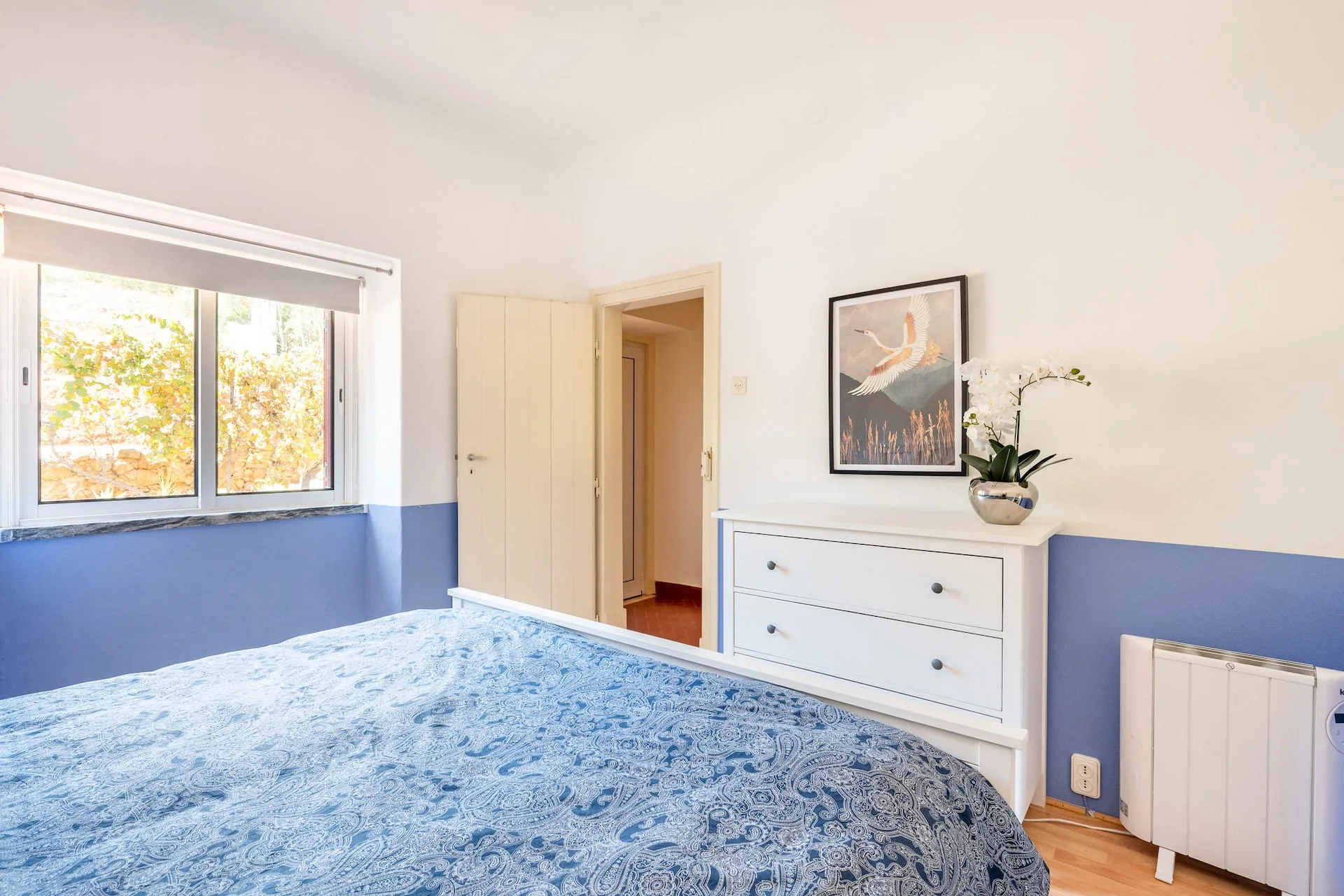 Bedroom with a double bed under a blue paisley bedspread, soft blue walls and a window looking out over the vines.