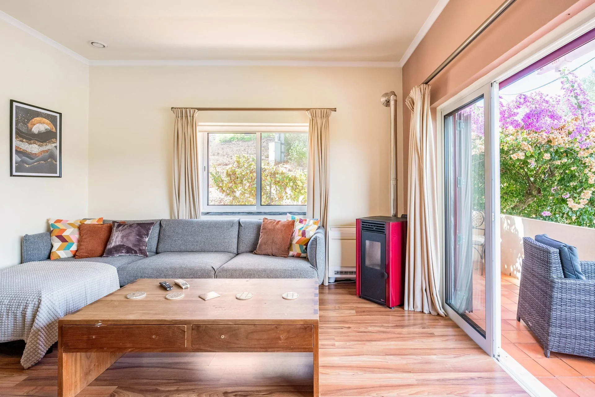 Living room with a grey corner sofa, bright cushions, a red wood-burning stove and sliding doors opening onto a bougainvillea-covered terrace.