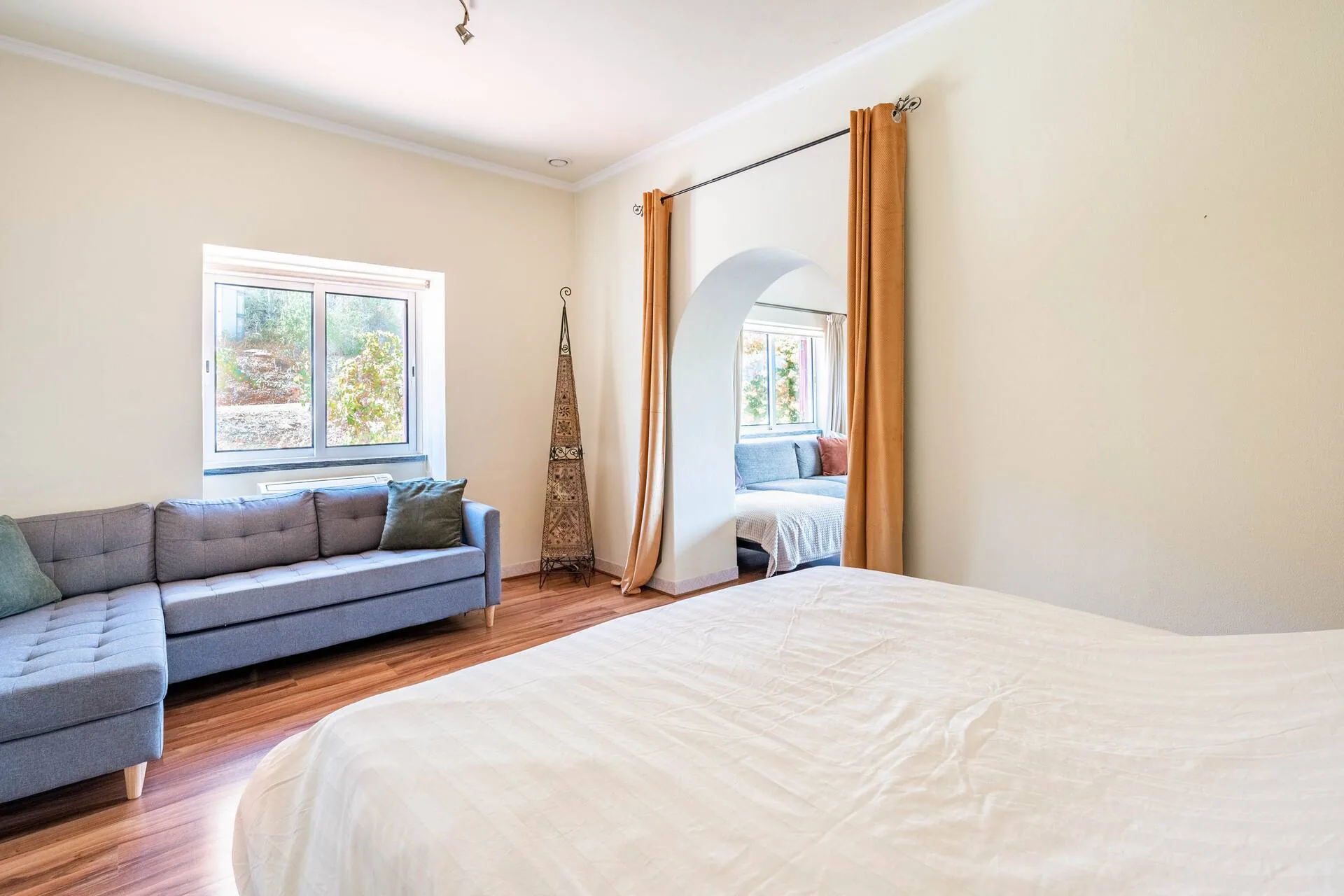 Spacious bedroom with cream bedding, its own sofa, and an archway framed by amber curtains leading to a second sitting area.
