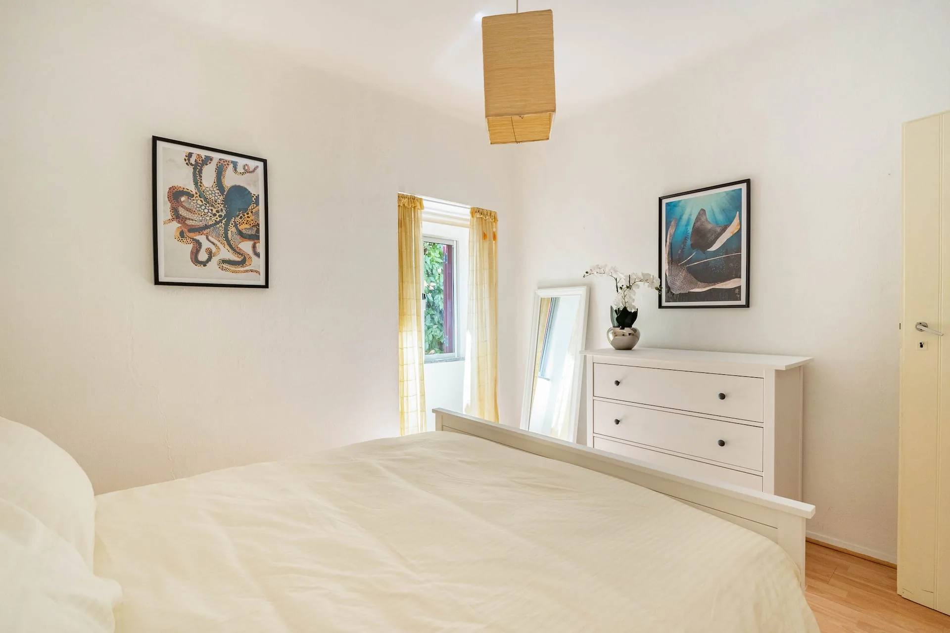 Bright bedroom with cream bedding, white walls, a straw pendant lamp and framed octopus and manta ray prints.